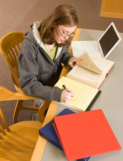 A female student is sitting at a desk in a library; she is taking notes from a large book. There are two binders on the table, along with a laptop computer. She is in the process of thesis editing.