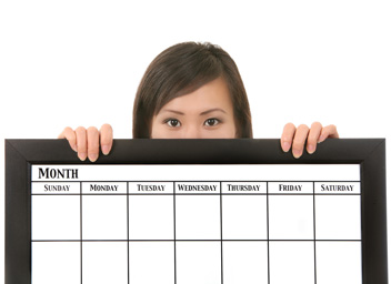 A woman is holding a large editorial calendar. The calendar covers the bottom half of her face and most of her body.