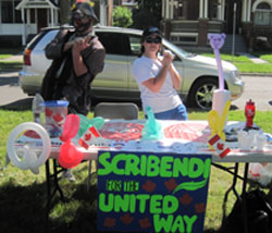 A clown and a young woman pose behind a table that has balloon animals and a sign on it said says, "Scribendi for the United Way."