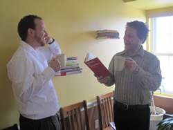 Two men are standing in a yellow room, each with a cup of coffee and one with a book. They are having an animanted conversation.