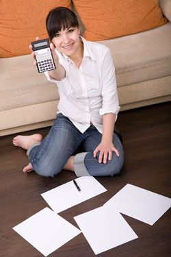 A young brunette woman is sitting on the floor of her house holding a calculator; there are pieces of paper that contain tax tips on the floor.